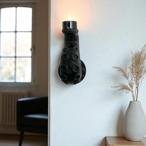 May include: A black octopus tentacle wall sconce with a candle-shaped light. The sconce is mounted on a white wall, with a round base and suction cup details. The light emits a warm glow, creating a unique decorative accent.