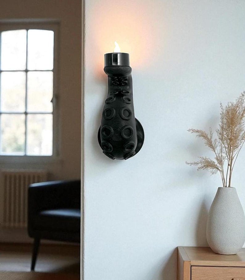May include: A black octopus tentacle wall sconce with a candle-shaped light. The sconce is mounted on a white wall, with a round base and suction cup details. The light emits a warm glow, creating a unique decorative accent.
