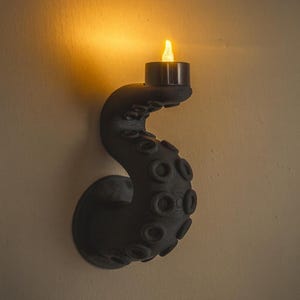 May include: A black wall-mounted candle holder shaped like an octopus tentacle. The tentacle has textured suckers and holds a small, battery-operated candle with a flickering yellow light. The candle holder is mounted on a beige wall.