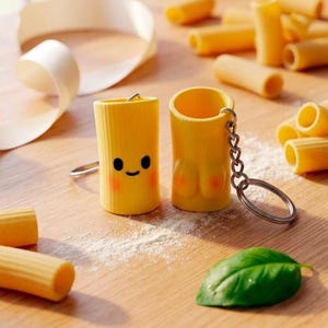 Op de afbeelding: Twee gele rigatoni pasta-vormige sleutelhangers met cartoongezichten en blush. Eén sleutelhanger heeft een lachend gezicht, de andere heeft een paar wangen. Op een houten oppervlak met pasta en een groen blad.