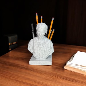 May include: A gray stone bust of a man with a toga, used as a pencil holder.  The bust is on a gray stone base and has several yellow pencils sticking out of the top.