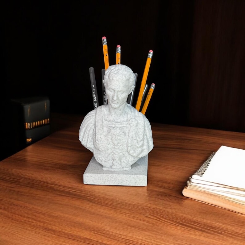 May include: A gray stone bust of a man with a toga, used as a pencil holder.  The bust is on a gray stone base and has several yellow pencils sticking out of the top.