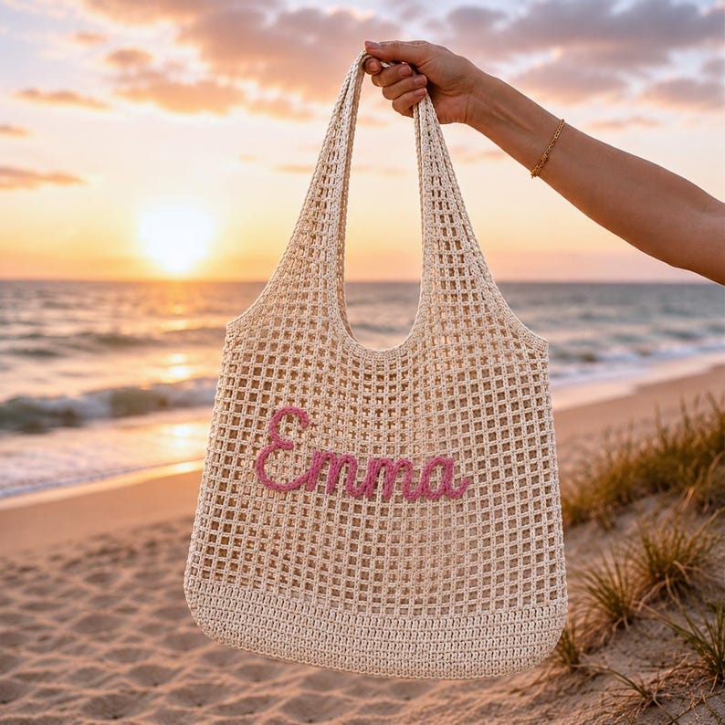 Personalized crochet beach tote bag with the name Emma in pink lettering, photographed at sunset by the ocean. Neutral cotton mesh shoulder bag with a coastal aesthetic, perfect for beach vacations, bridesmaid gifts, summer travel, pool days, and personalized gift ideas for women.