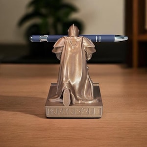 Puede incluir: Un portal&aacute;pices de color bronce con forma de caballero medieval, sosteniendo un bol&iacute;grafo azul. El caballero est&aacute; detallado con armadura y capa, de pie sobre una base rectangular. El bol&iacute;grafo es azul oscuro con detalles plateados.