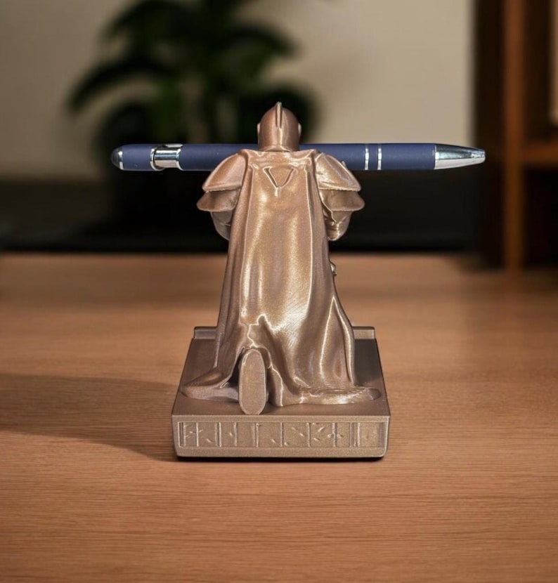 Puede incluir: Un portal&aacute;pices de color bronce con forma de caballero medieval, sosteniendo un bol&iacute;grafo azul. El caballero est&aacute; detallado con armadura y capa, de pie sobre una base rectangular. El bol&iacute;grafo es azul oscuro con detalles plateados.