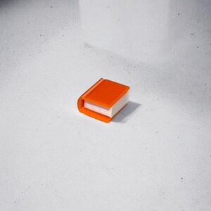 May include: A small orange book with white pages. The book is closed and sitting on a white surface.