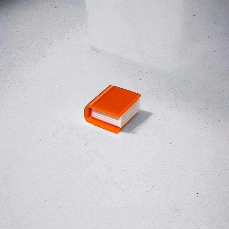 May include: A small orange book with white pages. The book is closed and sitting on a white surface.