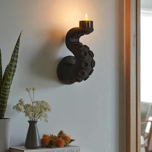 May include: A black octopus tentacle wall sconce with a lit candle. The sconce is mounted on a white wall. The tentacles have textured suckers. The candle emits a warm glow. Decorative items, including a vase of flowers, are on a nearby table.