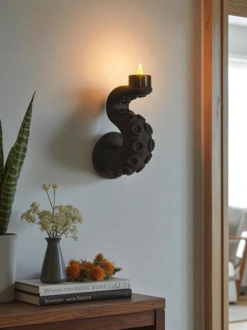 May include: A black octopus tentacle wall sconce with a lit candle. The sconce is mounted on a white wall. The tentacles have textured suckers. The candle emits a warm glow. Decorative items, including a vase of flowers, are on a nearby table.