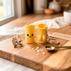 Peut inclure: Deux porte-clés cylindriques jaunes avec un design de dessin animé. L'un a un visage souriant, l'autre a une forme unique. Ils sont sur une planche à découper en bois avec une petite cuillère en laiton et du sel. L'étiquette indique "HECHO A MANO".