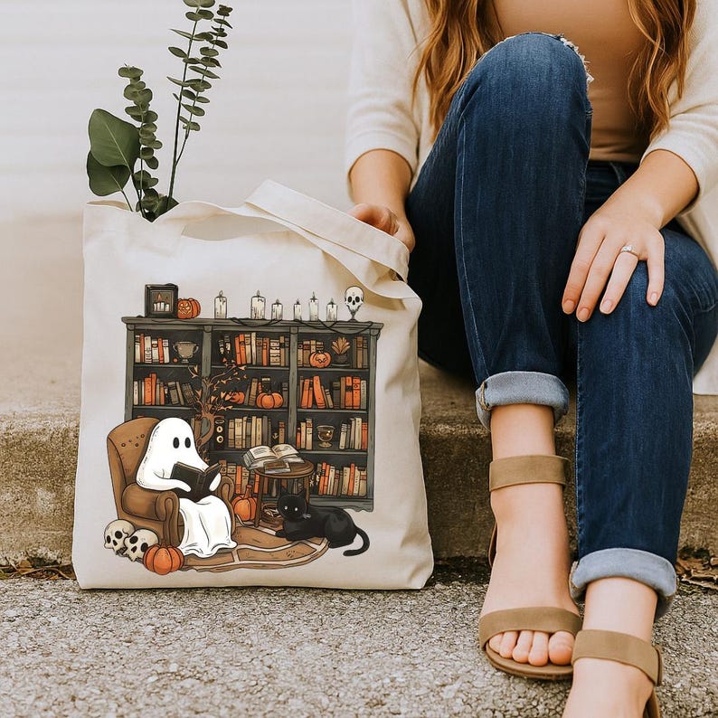 Op de afbeelding: Een witte tote bag met een griezelig Halloween-ontwerp. Een geest leest een boek in een bibliotheek met een zwarte kat en pompoenen.