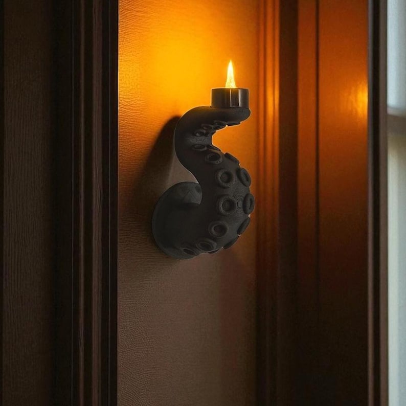 May include: A black wall-mounted candle holder shaped like an octopus tentacle. The tentacle curves upwards, holding a lit candle. The holder is mounted on a wooden surface, with warm lighting illuminating the scene.