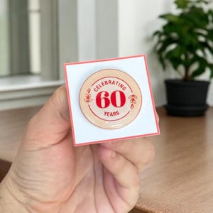 May include: A round pin with the text "Celebrating 60 Years" in red on a beige background. The pin is on a white card with a pink border. The pin has a floral design on the sides.