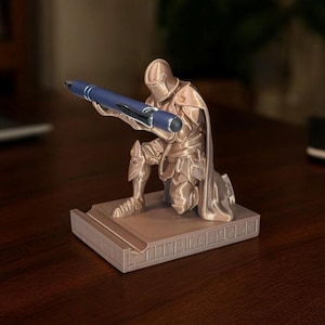 Puede incluir: Un portal&aacute;pices de color bronce con forma de caballero arrodillado sosteniendo un bol&iacute;grafo azul. El caballero est&aacute; detallado con armadura y una capa, descansando sobre una base rectangular. El bol&iacute;grafo se sujeta con ambas manos.