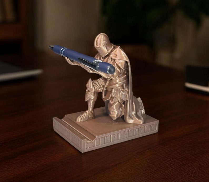 Puede incluir: Un portal&aacute;pices de color bronce con forma de caballero arrodillado sosteniendo un bol&iacute;grafo azul. El caballero est&aacute; detallado con armadura y una capa, descansando sobre una base rectangular. El bol&iacute;grafo se sujeta con ambas manos.