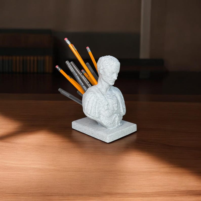 May include: A white plaster bust of a Roman soldier with a pencil holder in the back. The bust is on a square base and has a textured surface. There are several pencils in the holder.