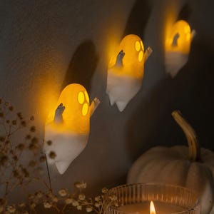 May include: Three small, white, ghost-shaped wall lights with glowing yellow interiors. Each ghost features cut-out eyes and an open mouth, with arms raised in a waving motion. The lights emit a warm, soft illumination against the wall surface.