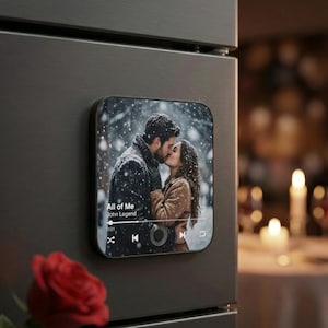 May include: A black square magnet with a photo of a couple kissing in the snow. The magnet displays a music player interface with the song "All of Me" by John Legend. A red rose is in the foreground.
