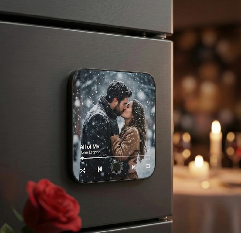 May include: A black square magnet with a photo of a couple kissing in the snow. The magnet displays a music player interface with the song "All of Me" by John Legend. A red rose is in the foreground.