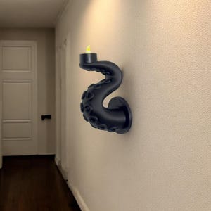 May include: A black wall-mounted candle holder shaped like an octopus tentacle. The tentacle curves upward, holding a small, black candle cup with a yellow LED flame. The holder is mounted on a textured, off-white wall.