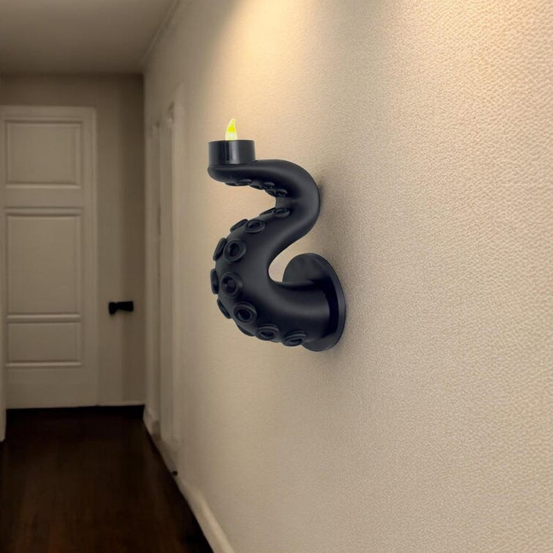 May include: A black wall-mounted candle holder shaped like an octopus tentacle. The tentacle curves upward, holding a small, black candle cup with a yellow LED flame. The holder is mounted on a textured, off-white wall.