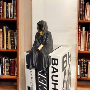 May include: A black hooded figure sits on a stack of books, holding a small orange book. The books are titled "BAUHAUS" and "The Kingdom", with the word "BAUHAUS" in large white letters.