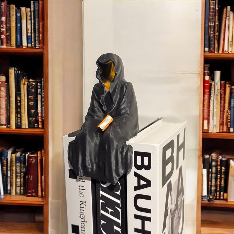 May include: A black hooded figure sits on a stack of books, holding a small orange book. The books are titled "BAUHAUS" and "The Kingdom", with the word "BAUHAUS" in large white letters.