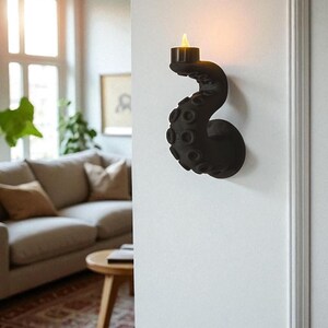 May include: Black octopus-shaped wall sconce holding a small candle with a flickering yellow flame. The sconce is mounted on a white wall. The design features a tentacle shape with suction cup details.