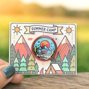 May include: A circular enamel badge with a red edge, depicting a camping scene with a cabin, trees, and a sunset. The badge is on a card with a banner reading "SUMMER CAMP" and illustrations of mountains, trees, and a tent.