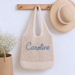 May include: A cream-coloured, open-weave tote bag with the name "Caroline" embroidered in blue. A straw sun hat with a white band hangs nearby. The bag is suspended from a wooden rack on a white wall. A woven basket with flowers is in the lower left.
