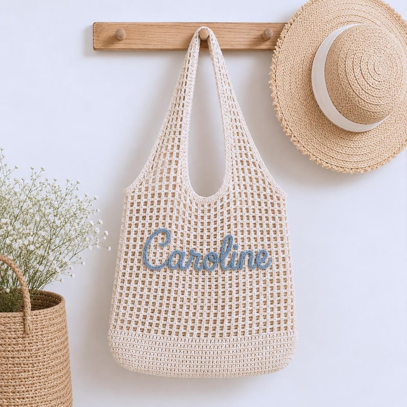 May include: A cream-coloured, open-weave tote bag with the name "Caroline" embroidered in blue. A straw sun hat with a white band hangs nearby. The bag is suspended from a wooden rack on a white wall. A woven basket with flowers is in the lower left.