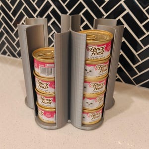 May include: A grey 3D-printed cat food can organiser holding several cans of Fancy Feast cat food. The organiser has a circular design with vertical slots to hold the cans. The cans are stacked inside the slots.