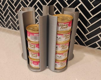May include: A gray 3D-printed cat food can organizer holding multiple cans of Fancy Feast cat food. The organizer has a circular design with vertical slots to hold the cans. The cans are stacked inside the slots.