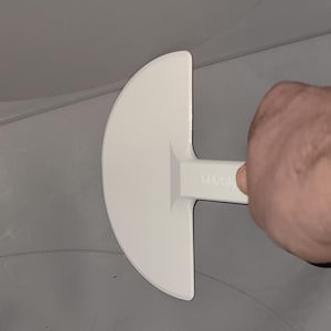 May include: A white plastic tool with a semi-circular head and a handle. The tool is held by a hand, and is positioned against a gray surface. The tool appears to be used for smoothing or spreading a material.