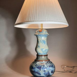 May include: A ceramic table lamp with a white pleated lampshade. The lamp base is a mix of blue, cream, and brown tones. The base has a bulbous bottom and a slender, decorative neck. The lamp is lit, casting a warm glow.
