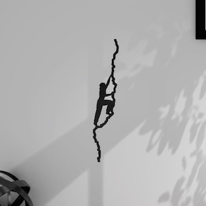 Op de afbeelding: Zwarte metalen wandsculptuur van een persoon die een rotswand beklimt. De sculptuur is bevestigd aan een witte muur.
