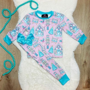 May include: Pink pajama set with a festive print featuring nutcrackers, gingerbread houses, and Christmas trees. The long-sleeved top and pants have blue trim. A large blue bow is attached to the pants. The pajama set is displayed on a white fluffy rug.