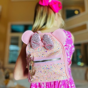 Peut inclure: Un sac à dos rose pailleté avec un grand nœud et des oreilles. Le sac à dos a une fermeture éclair et un tirette de fermeture éclair dorée.