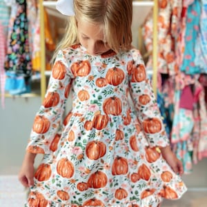 May include: A white long-sleeved dress with a pumpkin pattern. The dress features orange pumpkins, pink flowers, and green leaves. The dress has a ruffled hem and is suitable for fall. A white bow is in the hair.