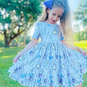 Può includere: Una bambina che indossa un vestito azzurro chiaro con sfondo bianco e una stampa di principesse Disney. Il vestito ha un orlo arricciato e maniche corte. La bambina indossa un fiocco blu nei capelli.