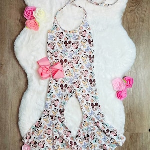 May include: A white halter-style jumpsuit with a print featuring Mickey Mouse and friends. The jumpsuit has flared legs and a pink bow. The background is a white fluffy rug with pink and yellow roses.