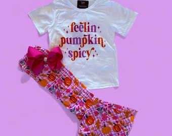 May include: White t-shirt with the text "feelin' pumpkin spicy" in orange and purple. Paired with bell-bottom pants featuring a pumpkin and floral pattern, and a large pink bow.