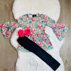 May include: A children's outfit featuring a long-sleeved top and black leggings. The top has a teal background with a pattern of nutcrackers and flowers, and bell sleeves. A large pink bow is included.