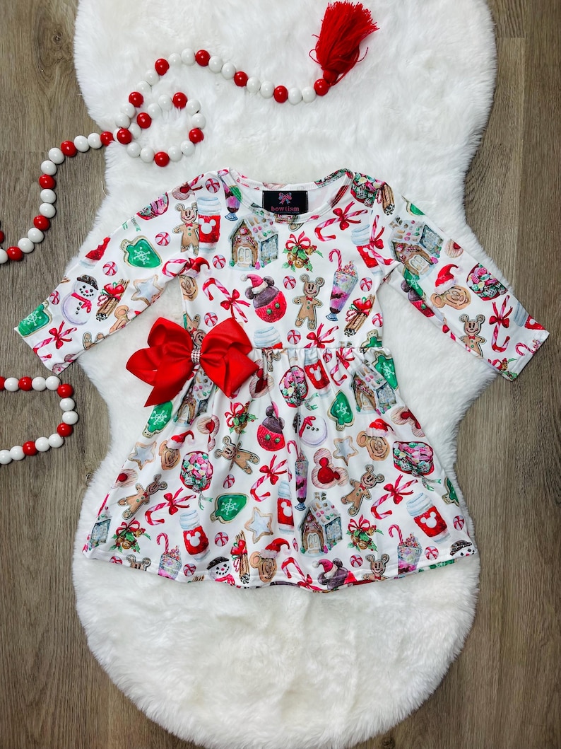 May include: White Christmas-themed dress with a red bow. The dress features a print of gingerbread men, candy canes, and other holiday treats.