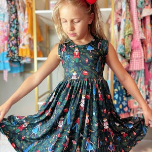 May include: A sleeveless dark teal dress featuring a festive Nutcracker print with red, blue, and white figures. The dress has a round neckline and a flared skirt. A large red bow is in the girl's hair.
