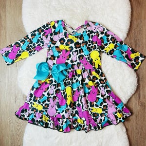 Peut inclure: Une robe blanche à imprimé de lapin rose, jaune, bleu et turquoise sur fond léopard. La robe a des manches longues et un ourlet à volants. Un nœud turquoise est noué autour de la taille.