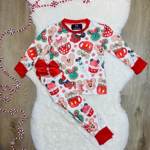 May include: White pajamas with a red trim. The pajamas feature a festive Christmas print with Mickey Mouse and Minnie Mouse characters, along with holiday treats like gingerbread cookies, hot chocolate, and candy canes.