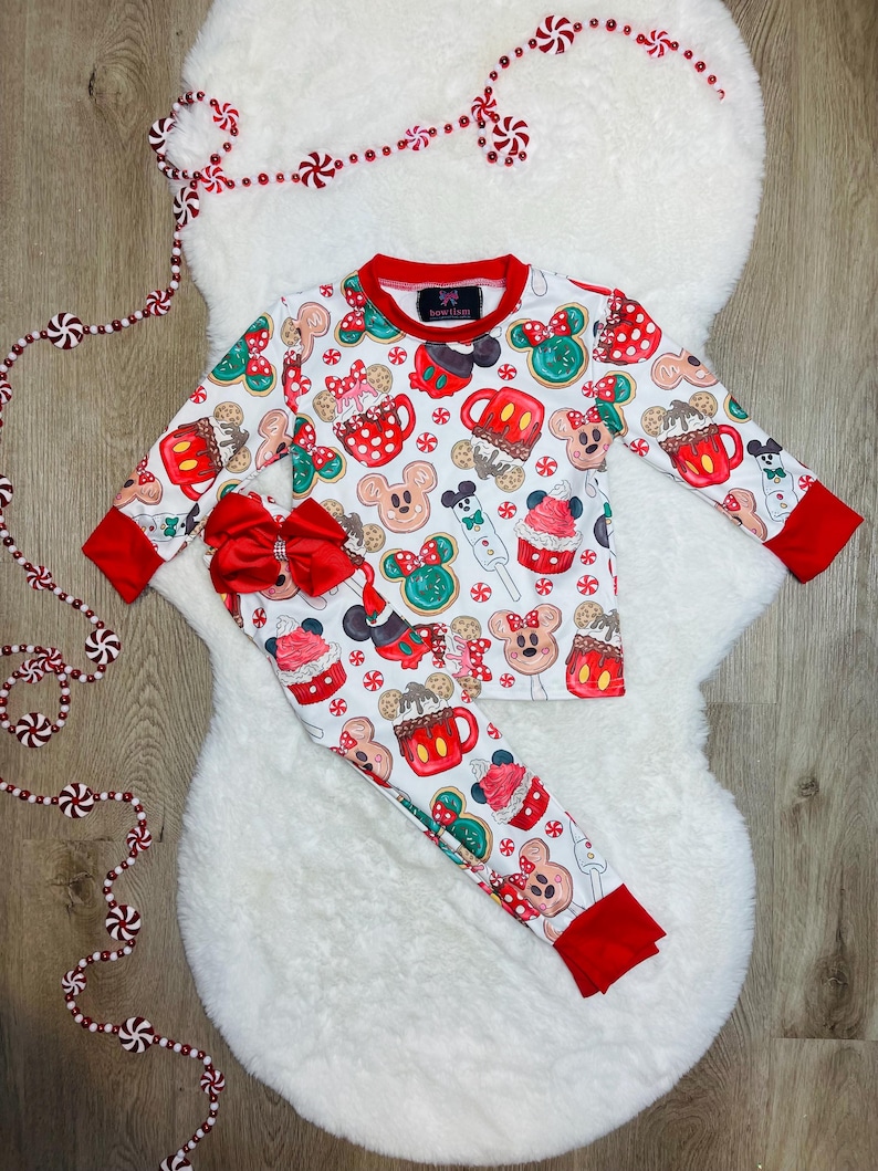 May include: White pajamas with a red trim. The pajamas feature a festive Christmas print with Mickey Mouse and Minnie Mouse characters, along with holiday treats like gingerbread cookies, hot chocolate, and candy canes.