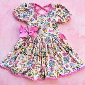 May include: A beige dress with short puff sleeves and a pink trim, featuring a repeating pattern of yellow school buses, pencils, rainbows, books, and flowers. A large pink bow is attached to the waist, and the back has a crisscross strap design.