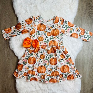 May include: A white dress with a pumpkin and floral print, featuring long sleeves and a ruffled hem. An orange bow is attached at the waist. The dress is designed for children and is suitable for fall or Halloween.